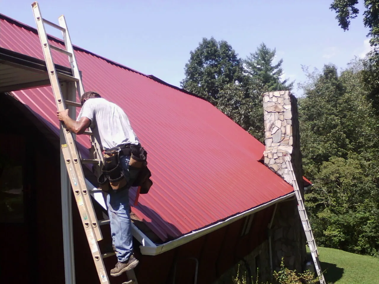 Roofer on a ladder beside a red metal roof and stone chimney — Soffit & Fascia Repair by Roofing Brothers in Lyndonville